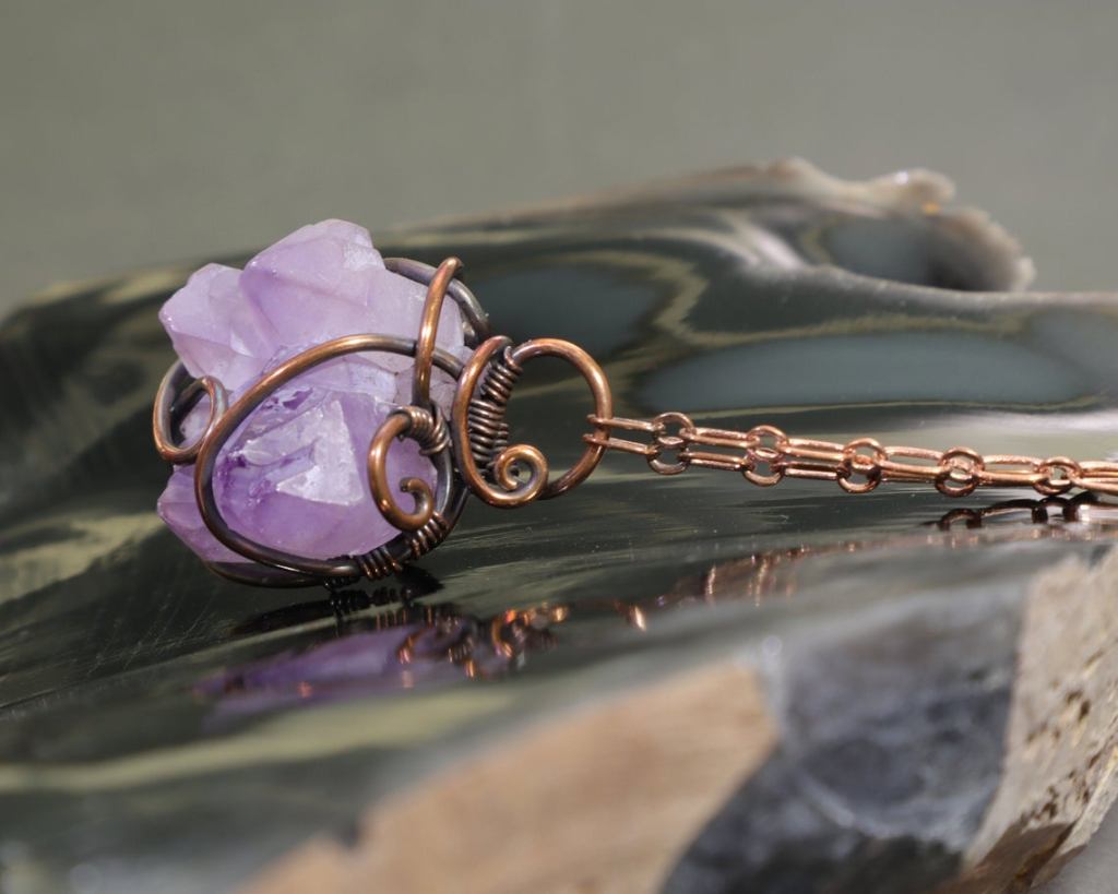 Raw elestial amethyst crystal wire-wrapped with copper wire on a copper chain and sitting on a piece of broken obsidian