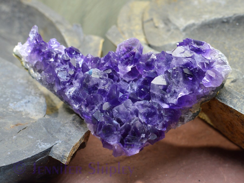 A dark purple Uruguayan amethyst crystal cluster on matrix balanced on two rocks with rocks in the background.