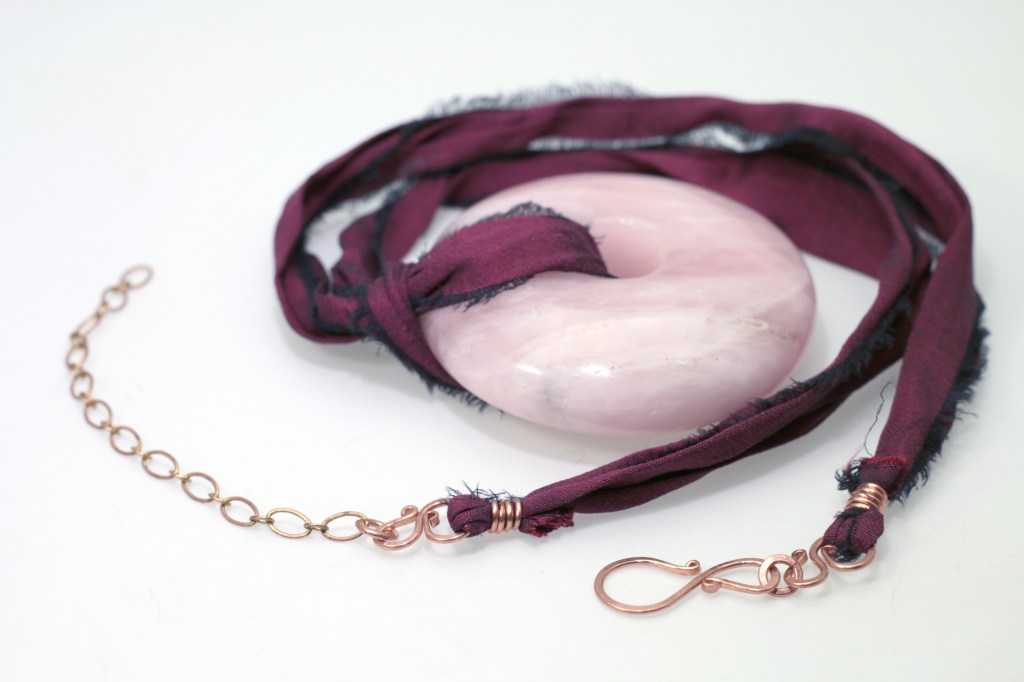 A rose quartz crystal donut on a recycled sari silk necklace with hand forged and hammered copper clasps and an extender chain