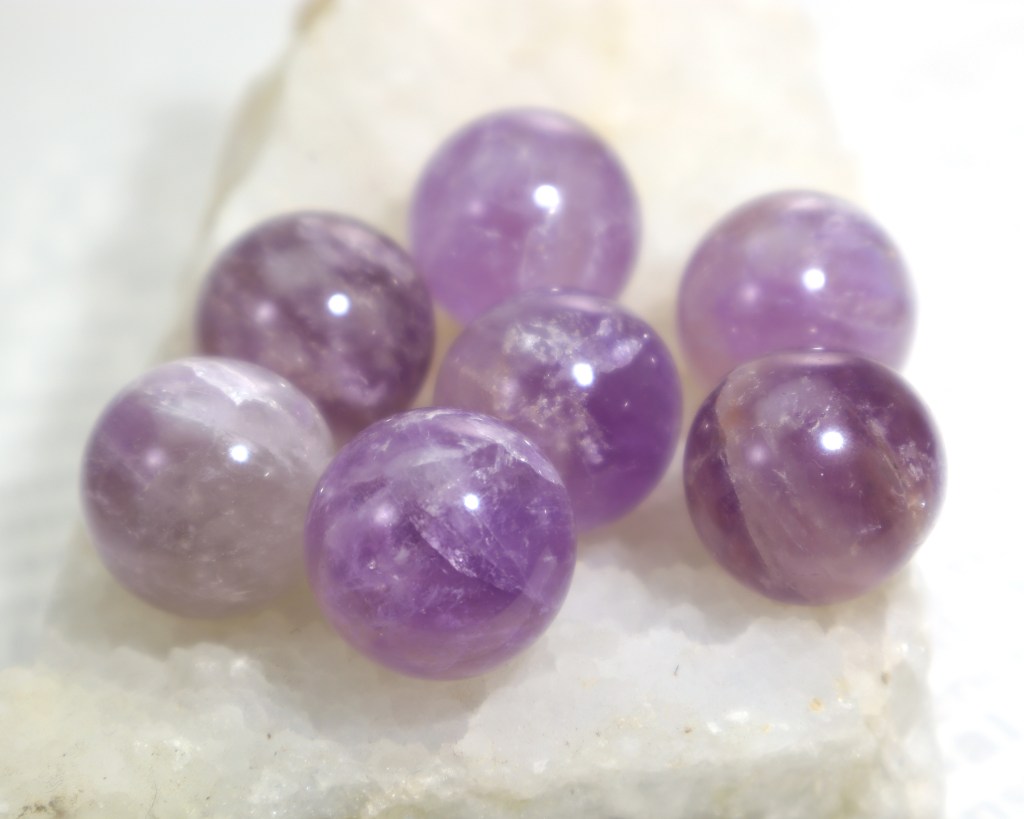 A collection of purple amethyst crystal spheres with a various violet shades and inclusions on a white quartz cluster in front of a white background